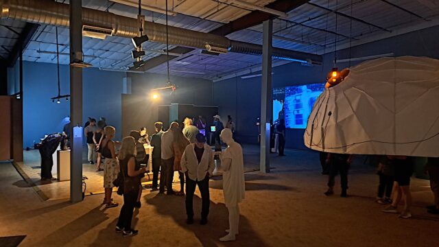 People in a dark-ish warehouse like room surrounded by new media art