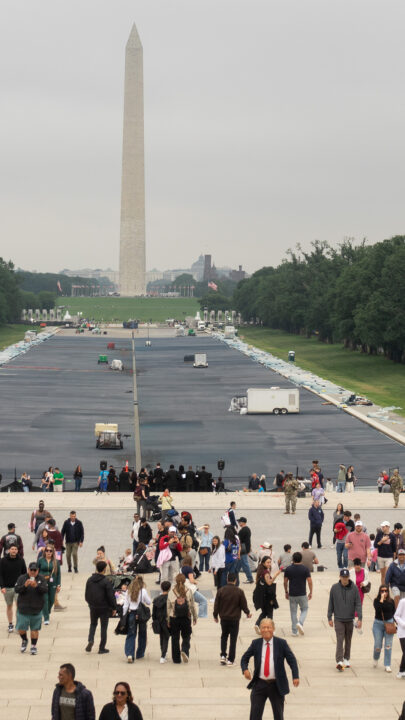 View of the empty reflecting pool being painted blue with the Washington Monument in the background. A person in a POTUS mask can be seen on the steps of the Lincoln Memorial in the foreground.