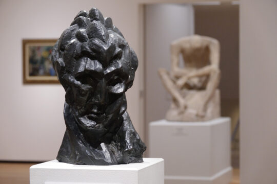 Close up of dark head sculpture by Picasso with a statue of a sitting man in the background.