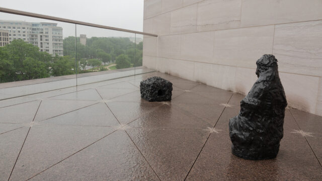 A small statue of a figure looking at a box made of the same material. They are located on a roof terrace with a high-rise and some trees in the background